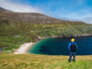 Wall Mural - A man in a blue jacket stands on a hill overlooking a beach and a ocean. The sky is clear and the sun is shining, creating a peaceful and serene atmosphere. Keem bay and beach Ireland. Travel concept