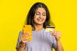 © Andrii Iemelianenko - Happy Indian young woman using credit bank card and smartphone while transferring spending money, purchases online shopping, ordering food delivery. Arabian Hindu girl isolated on yellow background