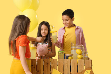 Wall Mural - Cute little children with piggy bank selling lemonade at stand on yellow background