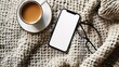 © naphat - A photo of a modern smartphone with a blank screen lying flat on a textured fabric background. The phone is positioned near a cup of coffee and a pair of stylish eyeglasses.