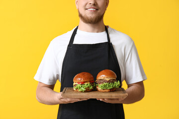 Wall Mural - Young man in apron holding board with tasty burgers on yellow background