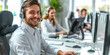 © Duka Mer - A young male call center agent, wearing a headset, works in an office with his team of customer service workers in the background.