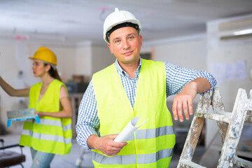 Wall Mural - Portrait of positive foreman standing at stepladder in apartment. Woman painting wall in background, renovation works in house.