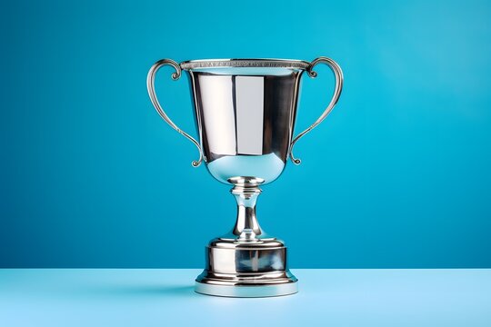 Shiny silver trophy with an elegant base, standing against a blue background. The cup is adorned with intricate designs and features two hands holding the handle at its top. It has been decorated.