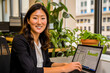 © apratim - Smiling businesswoman working on a laptop in a modern office with plants and large windows in the background.