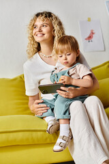 Wall Mural - A curly-haired mother sits on a couch, lovingly embracing her toddler daughter at home.