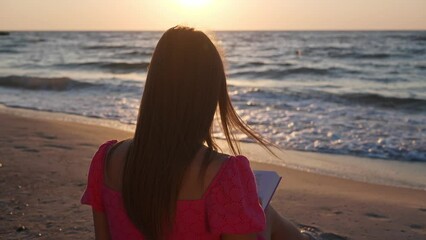 Wall Mural - Woman reading book on beach by sea in morning, back view. Camera moving around girl