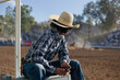 © Connect Images - A person is sitting on a rail, wearing a cowboy hat and plaid shirt, looking at a mobile phone with a rodeo arena in the background.