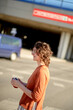 © Connect Images - A person with curly hair is smiling and standing in a sunlit area, possibly a parking lot, with a clear blue sky above and a building with red text in the background.