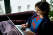 © Connect Images - A person is sitting on a couch working on a laptop, dressed in a blue sweater with a red pattern, with a blanket over their legs.