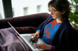 © Connect Images - A woman is sitting on a purple couch using a laptop. She appears focused on her work, with natural light illuminating the room from a window beside her.