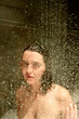 © Connect Images - A young woman stands in a shower with water droplets cascading down, wet hair plastered to their head, and a neutral expression on their face.