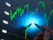 © Connect Images - A person's hand holds a pen, pointing at a bright screen displaying a stock market graph with upward trends in green and blue tones.