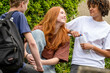 © Connect Images - Three friends are engaged in a lively conversation outdoors. A redhead woman is laughing with a woman holding a water bottle, while a man with a backpack looks on.