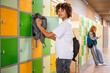 © Connect Images - A young adult with curly hair is putting a jacket into a yellow locker in a school hallway, while another person is using a phone in the background.