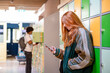© Connect Images - A young girl with red hair is focused on her smartphone in front of green lockers, with a young boy in the background carrying a backpack.