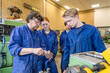 © Connect Images - A group of four people dressed in blue work overalls attentively watches as one of them demonstrates something with a metal part in a workshop environment.