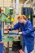 © Connect Images - A woman in a blue work coat operates machinery in a workshop, focusing intently on her task.