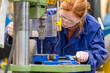 © Image Source - A woman with red hair is operating a drill press in an industrial setting, focused on her work wearing safety goggles and a blue work coat.