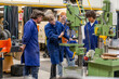 © Connect Images - Four individuals in blue work overalls are engaged in an activity in a workshop with various machinery and equipment around them.