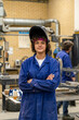 © Connect Images - A young adult wearing a blue welding jacket and a protective helmet stands confidently with crossed arms in a workshop environment, with equipment and another person in the background.