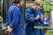 © Connect Images - Two workers in blue overalls are operating machinery in an industrial setting, with one observing while the other concentrates on the task.