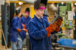 © Connect Images - A worker in protective gear examines a metal part in an industrial setting, with colleagues working in the background.