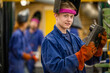 © Connect Images - A young worker in a blue jumpsuit and orange gloves is holding a hammer in a workshop, with two colleagues in the background.