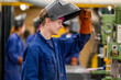 © Connect Images - A smiling young man wearing a blue work uniform is lifting his welding mask with a gloved hand in an industrial setting.