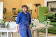© Connect Images - A man in a blue work uniform stands in a workshop with machinery and workbenches around him.