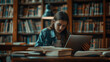 © nopommajun - A female student focusing intently on her laptop screen, surrounded by open books in a library setting