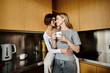 © Bliss - Two young lesbian women stand together in a hotel kitchen, chatting over coffee cups.