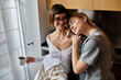 © Bliss - Two women, a young lesbian couple, stand in a cozy hotel room kitchen, each with a coffee cup in hand.