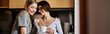 © Bliss - Two women, a young lesbian couple, stand in a hotel room kitchen, holding coffee and engaged in conversation.