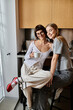 © Bliss - A young lesbian couple sitting closely, enjoying coffee in a hotel room kitchen.