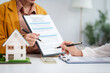 © NanSan - Asian businesswoman discusses home purchase contracts with clients at her desk, focusing on loans, mortgages, and other financial aspects of real estate transactions.