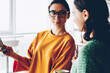 © BullRun - Beautiful female designer in eyeglasses showing information on smartphone to skilled student.Two entrepreneurs discussing post in social network standing together in office interior