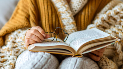 Close-up of relaxed person reading a book, holding glasses, wrapped in cozy blanket. Calm and comfortable atmosphere.