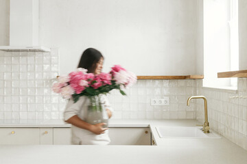 Blurred woman arranging peonies in vase on background of minimal white kitchen in new modern home. Motion image of young female decorating house after renovation, housekeeping