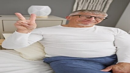 Canvas Print - Cheerful middle age, grey-haired man wearing pyjamas, smiling and confidently flashing a victory sign, fingers forming v for win, looking at the camera in his cozy bedroom.