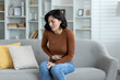 © Tetiana - A young woman grimacing in pain is sitting on the sofa at home and holding her stomach with her hand, feeling severe pain