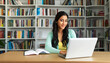 © Carmen - Young Hispanic woman student working at laptop with book shelves in background, copy space
