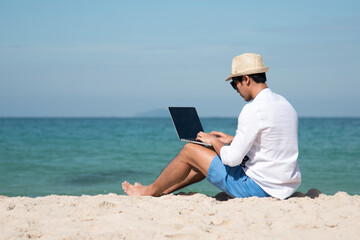 Wall Mural - Man traveler use laptop on summer beach blue sky. Asian man casual blue short pants white shirt relax sitting summer beach. Man typing laptop computer holiday business trip freelance work on island