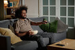 © AnnaStills - Young biracial guy sitting relaxed on couch in living room switching TV channels, copy space