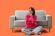 © Pixel-Shot - Thoughtful young woman sitting near sofa against orange background