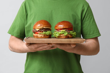 Wall Mural - Man holding board with tasty burgers on grey background