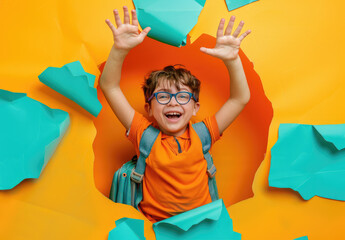 happy little boy is breaking through the yellow background wall with his hands up and wearing glasses, carrying a schoolbag and happily smiling at us while doing funny things.
