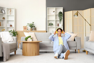 Canvas Print - Young African-American woman in headphones sitting beside sofa at home