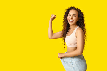 Canvas Print - Happy young African-American woman in loose jeans showing muscles on yellow background. Weight loss concept