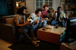 © AnnaStills - Group of joyful friends sitting in living room having fun watching soccer game match on TV late in evening